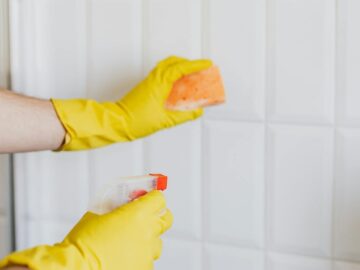 A person wearing yellow gloves cleans bathroom tiles with a sponge and spray bottle.